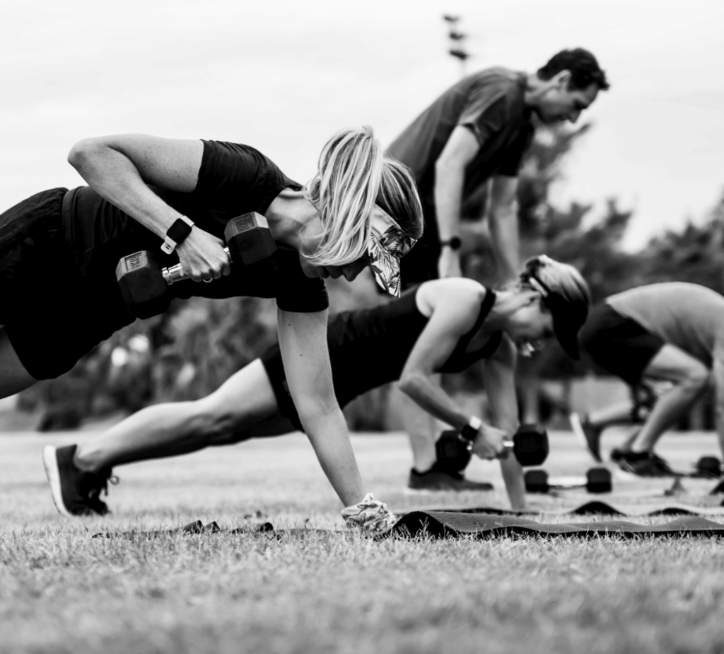 Outdoor group fitness class: participants in push-up position with dumbbells on mats, performing weighted push-ups in a park, black-and-white photo.