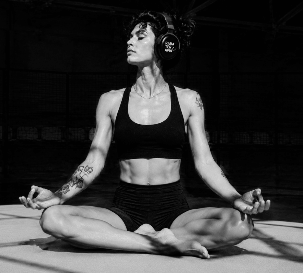 Woman meditating in a cross-legged lotus pose on a mat, wearing a black sports bra and shorts with over-ear headphones.
