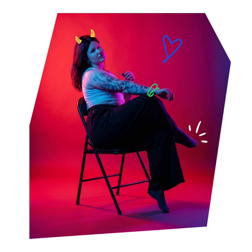 Portrait of a tattooed woman sitting on a black folding chair in a red-to-blue neon-lit studio, with playful neon doodles (horns, heart, and accents) around her.
